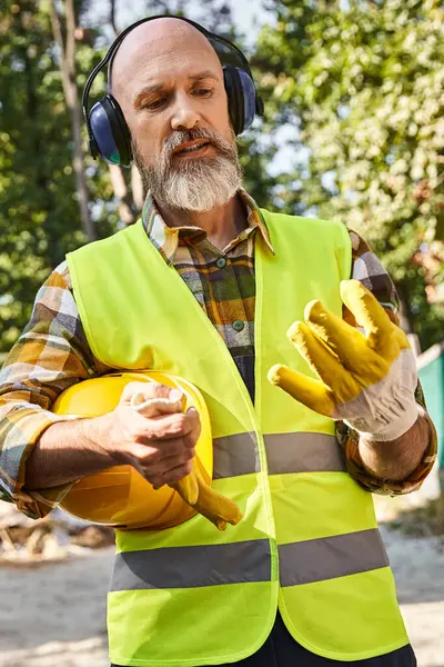 Constructeur de chalet travailleur en gants de sécurité et gilet posant avec casque et casque sur place — Photo de stock