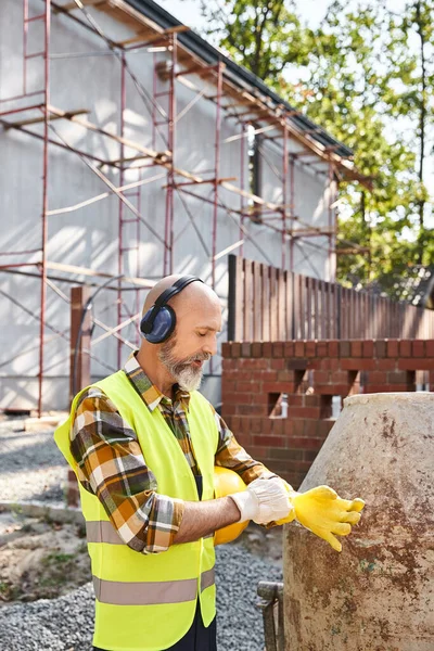 Trabalhador construtor de casa de campo em luvas de segurança e colete posando com fones de ouvido e capacete no local — Fotografia de Stock
