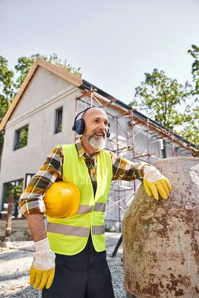 Alegre constructor de casa de campo en guantes de seguridad y chaleco posando con auriculares y casco en el sitio - foto de stock
