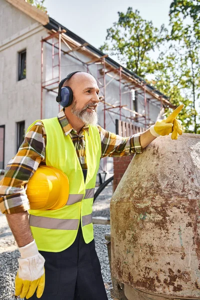 Cheerful cottage builder in safety gloves and vest posing with headphones and helmet on site — Stock Photo
