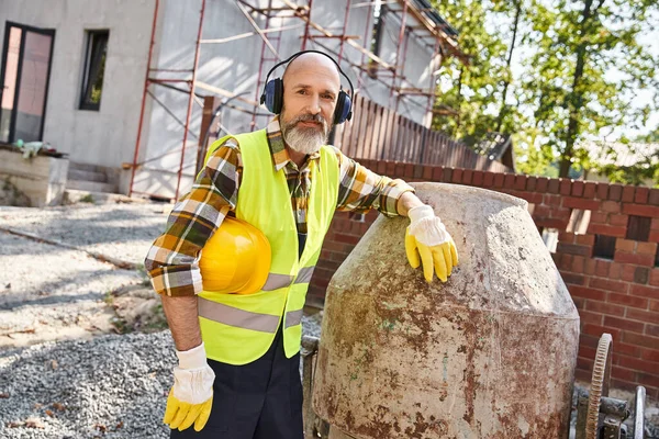 Весёлый строитель коттеджей в защитных перчатках и жилете с наушниками и шлемом, улыбающийся в камеру — стоковое фото