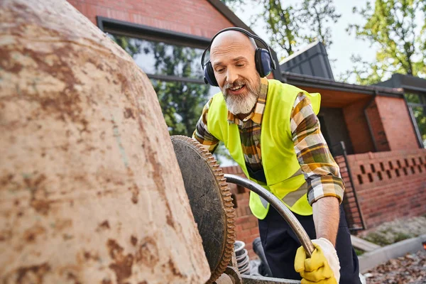 Handsome cheerful man in safety gloves and headphones working with circular saw, cottage builder — Stock Photo