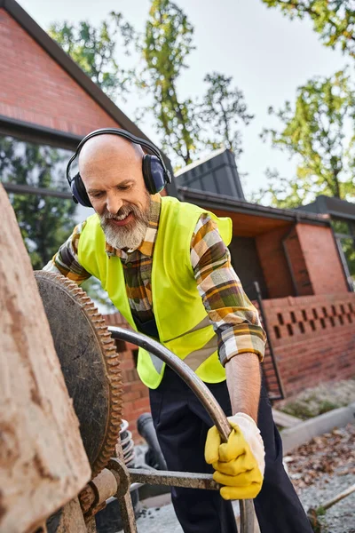 Handsome cheerful man in safety gloves and headphones working with circular saw, cottage builder — Stock Photo