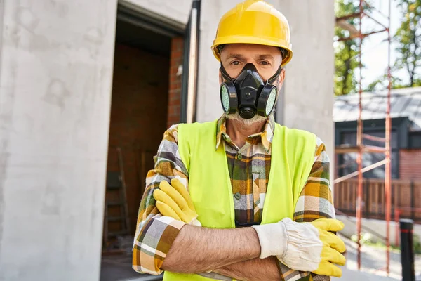 Atractivo hombre en guantes de seguridad y casco con máscara de polvo y mirando a la cámara, constructor de cabañas - foto de stock