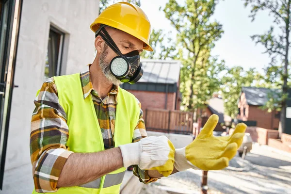 Dedicated handsome cottage builder in safety gloves and dust mask preparing to his work on site — Stock Photo