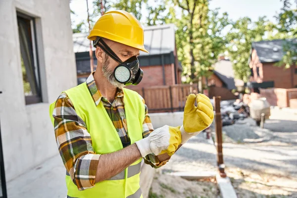 Dedicated handsome cottage builder in safety gloves and dust mask preparing to his work on site — Stock Photo