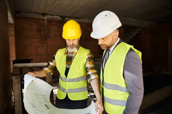 Two handsome cottage builders in safety vests and helmets discussing blueprint on construction site — Stock Photo