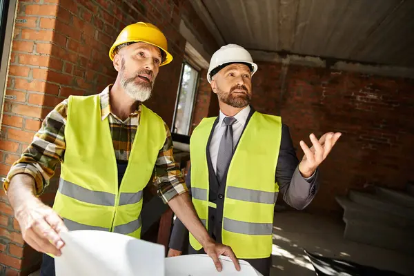 Jolly cottage builder and busimessman with blueprint discussing their work on construction site — Stock Photo