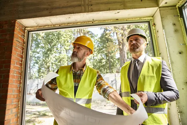 Handsome bearded businessman and cottage builder with safety gear working with blueprint on site — Stock Photo