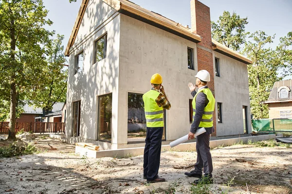 Hardworking cottage builder and architect holding blueprint and discussing construction site — Stock Photo