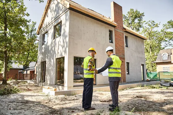 Hardworking cottage builder and architect holding blueprint and discussing construction site — Stock Photo