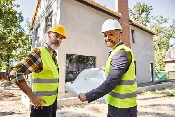 Cheerful builder and architect with safety gear holding blueprint and discussing construction site — Stock Photo