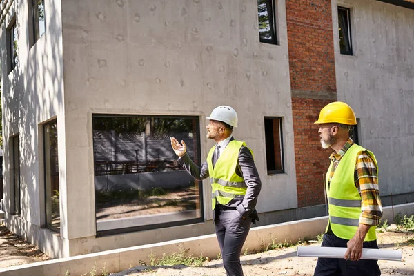 Commited architect and builder in safety vests holding blueprint and discussing construction process — Stock Photo