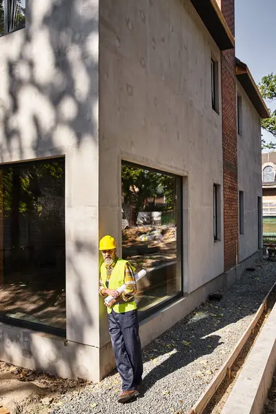 Good looking dedicated cottage builder in safety gear with blueprint and posing on construction site — Stock Photo