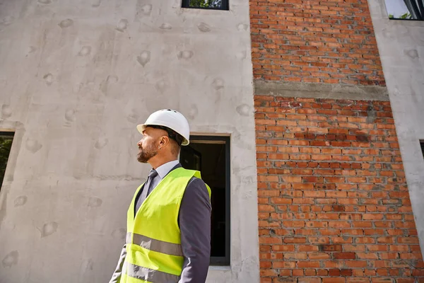 Homme d'affaires dévoué en gilet de sécurité et casque posant sur le chantier et regardant loin — Photo de stock