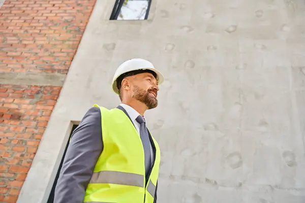 Alegre arquitecto dedicado en traje y chaleco de seguridad y casco sonriendo mientras que en el sitio de construcción - foto de stock