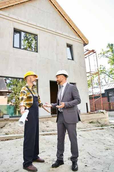 Jolly cottage builder and architect in suit and overalls with blueprint discussing construction — Stock Photo