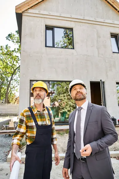 Dedicated architect and builder in suit and overalls with blueprint discussing construction process — Stock Photo