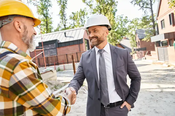 Joyous handsome architect and builder in overalls and suit with safety helmets shaking hands happily — Stock Photo