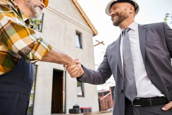 Cheerful handsome architect and builder in overalls and suit with helmets shaking hands happily — Stock Photo