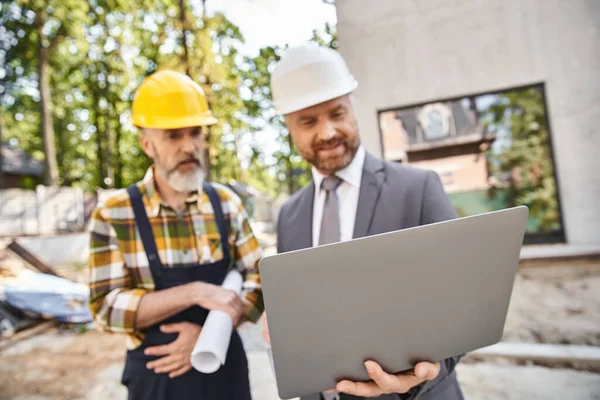 Focus on laptop in hands of blurred architect and builder in safety helmets on construction site — Stock Photo
