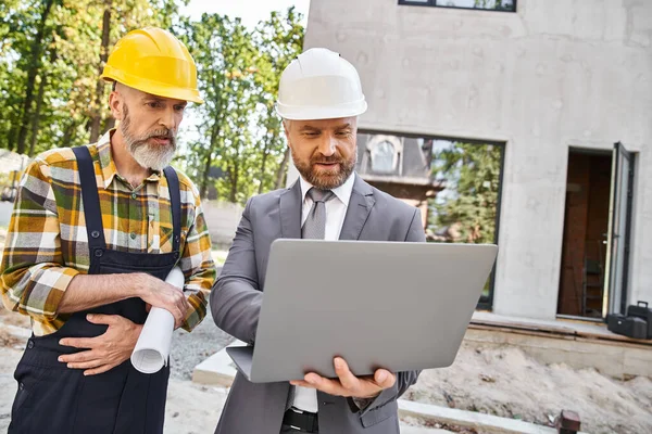 Arquitecto guapo y constructor en overoles y traje de trabajo con el ordenador portátil en el sitio de construcción - foto de stock