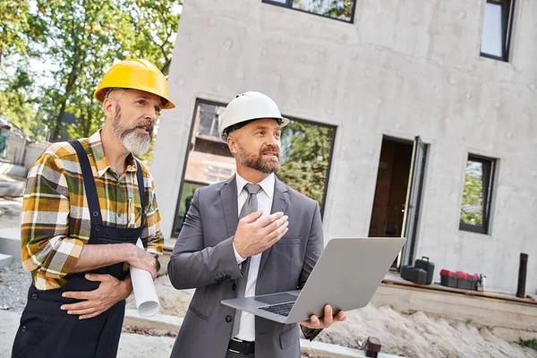 Appealing architect and builder in overalls and suit working with laptop on construction site — Stock Photo