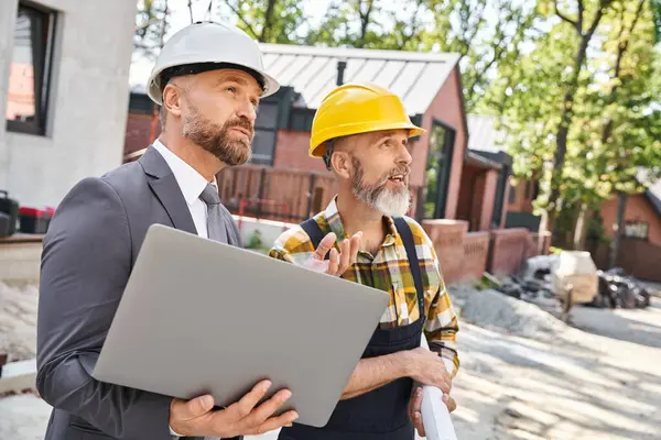Joyous handsome architect and builder in overalls and suit working with laptop on construction site — Stock Photo