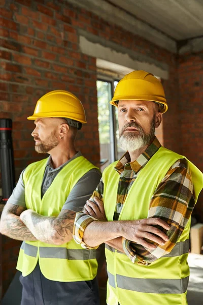 Dedicated bearded cottage builders in safety helmets and vests posing together with arms crossed — Stock Photo