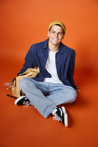 Attractive young man in yellow hat sitting cross-legs with backpack against terracotta background — Stock Photo