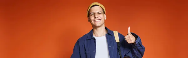 Banner of happy young guy in hat standing with backpack and showing like on terracotta background — Stock Photo