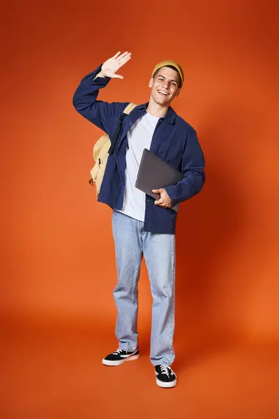 Junger Mann in lässigem Outfit und gelbem Hut mit Rucksack, Laptop in der Hand und Hallo sagen — Stockfoto