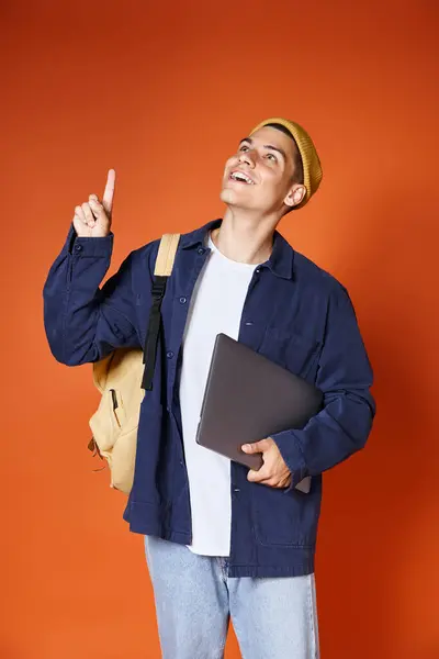 Hombre joven en sombrero amarillo con mochila que se le ocurrió la idea y sostener el ordenador portátil en fondo de terracota - foto de stock