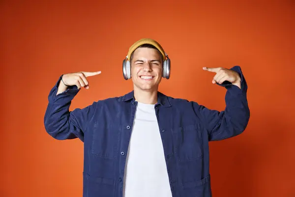 Fröhlicher junger Mann mit gelbem Hut zeigt mit den Fingern auf Kopfhörer vor Terrakottahintergrund — Stockfoto