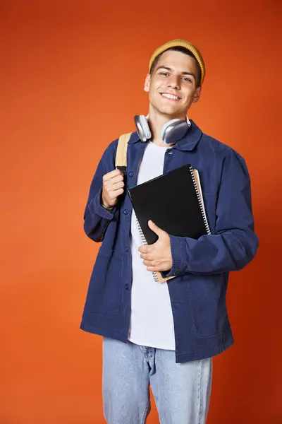 Charming student in headphones and yellow hat holding backpack and notes on terracotta background — Stock Photo