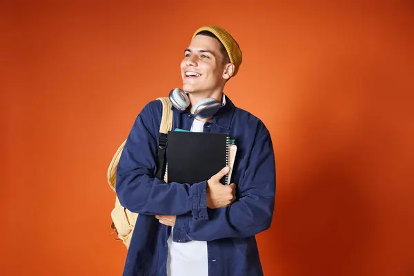 Jovem estudante sorrindo em fones de ouvido e chapéu amarelo segurando mochila e abraçando notas — Fotografia de Stock