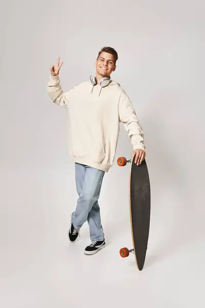 Handsome student in light hoodie and headphones standing with skateboard and showing sign peace — Stock Photo