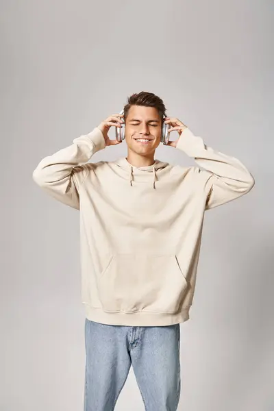 Attractive young man in light hoodie listening to music with closed eyes on light background — Stock Photo