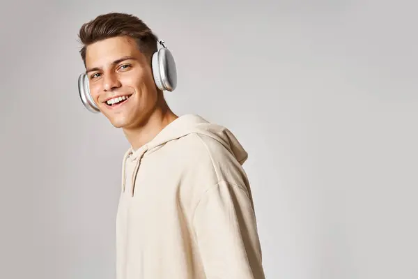Attractive young guy with brown hair in headphones smiling to camera against light background — Stock Photo