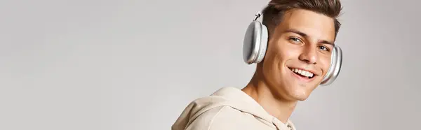 Horizontal shot of young guy with brown hair in headphones smiling to camera on light background — Stock Photo