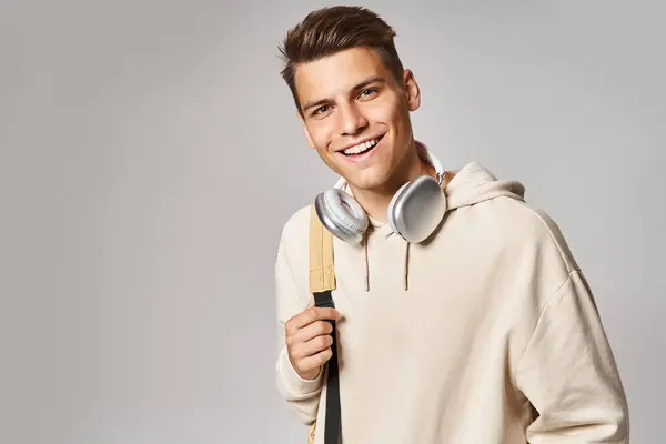 Jovem estudante atraente em fones de ouvido e roupa casual com mochila contra fundo cinza — Fotografia de Stock