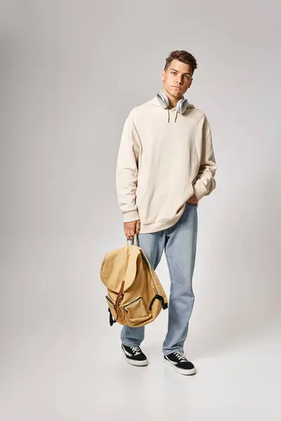 Estudiante guapo en auriculares y ropa casual caminando con mochila contra fondo gris - foto de stock