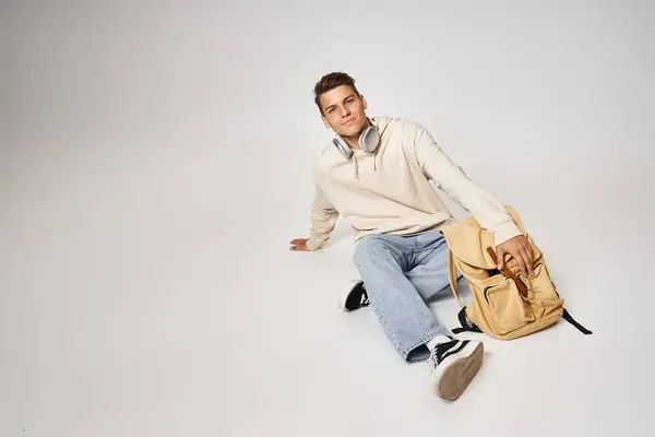 Handsome young man in headphones and casual outfit sitting with backpack on grey background — Stock Photo
