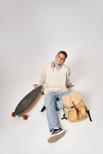 Atractivo estudiante en auriculares y atuendo casual sentado con mochila y monopatín - foto de stock