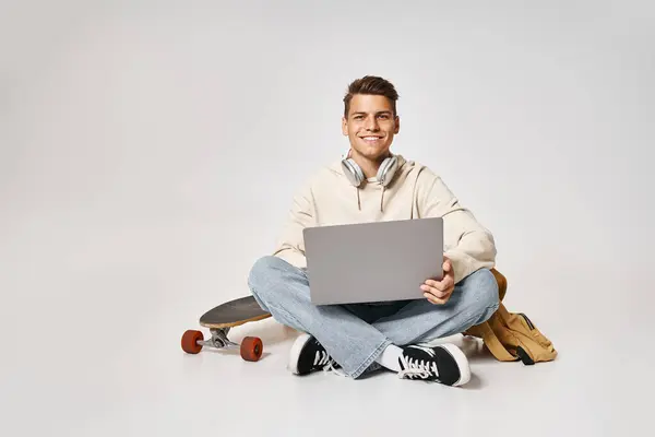 Smiling young student in headphones networking to laptop and sitting with backpack and skateboard — Stock Photo
