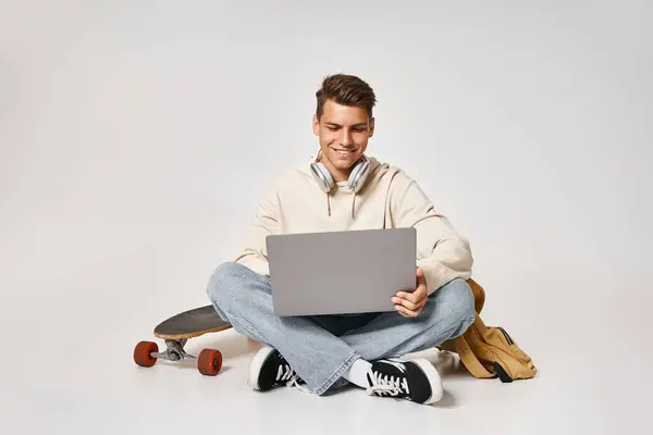 Estudiante inteligente en la red de auriculares a la computadora portátil y sentado con mochila y monopatín - foto de stock