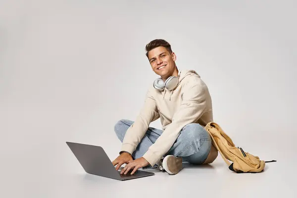 Веселий хлопець в навушниках мережа до ноутбука і сидить з рюкзаком на сірому фоні — стокове фото