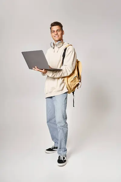 Estudiante joven en los auriculares de pie con la mochila y la red a la computadora portátil contra el fondo gris - foto de stock