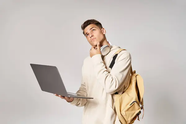 Hombre reflexivo en los auriculares con la red de la mochila a la computadora portátil y mirando hacia arriba en el fondo gris - foto de stock