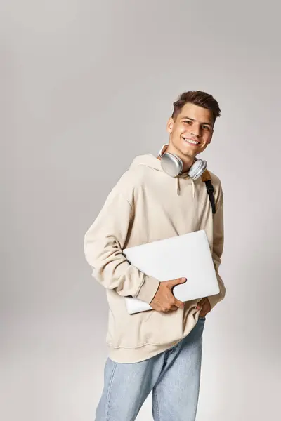 Handsome student in his 20s with laptop and headphones walking against grey  background — Stock Photo